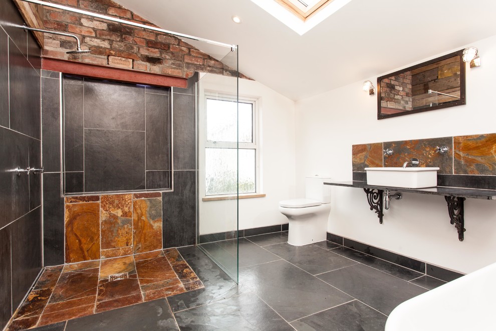 Industriell inredning av ett mellanstort badrum, med ett fristående badkar, en öppen dusch, svart kakel, stenkakel, vita väggar, skiffergolv, ett fristående handfat, en toalettstol med separat cisternkåpa och med dusch som är öppen