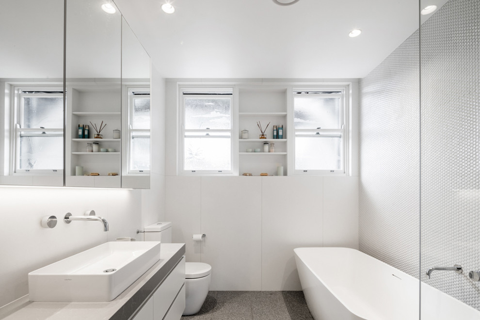 На фото: главная ванная комната в современном стиле с белыми стенами, плоскими фасадами, белыми фасадами, отдельно стоящей ванной, белой плиткой, плиткой мозаикой, настольной раковиной, серым полом и белой столешницей