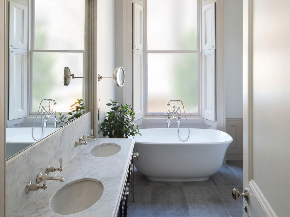 ロンドンにあるトラディショナルスタイルのおしゃれなマスターバスルーム (置き型浴槽、グレーのタイル、磁器タイル、グレーの壁、磁器タイルの床、一体型シンク、大理石の洗面台) の写真