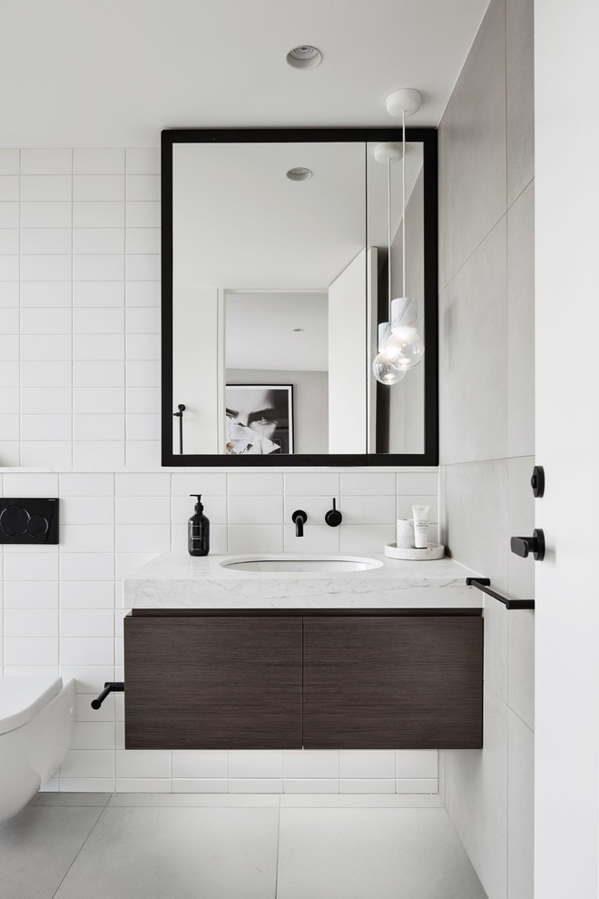 Mid-sized trendy white tile and ceramic tile ceramic tile and gray floor bathroom photo in Melbourne with furniture-like cabinets, brown cabinets, a wall-mount toilet, white walls, an undermount sink and marble countertops