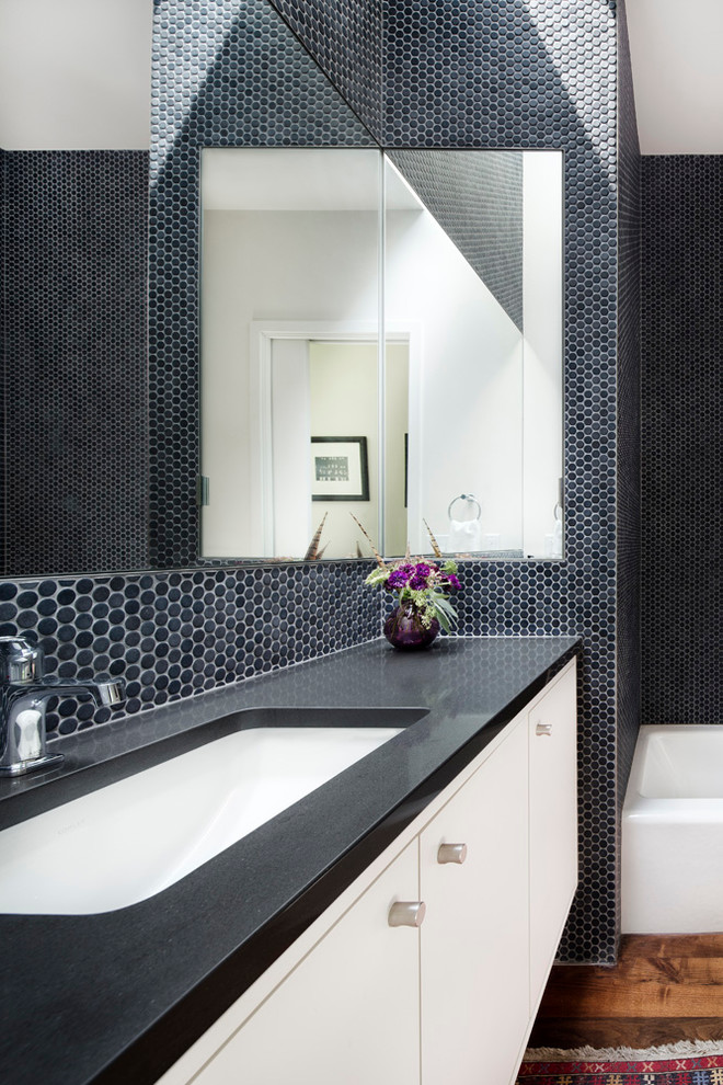 Cette image montre une salle d'eau design de taille moyenne avec un lavabo encastré, un placard à porte plane, des portes de placard blanches, un plan de toilette en surface solide, une baignoire en alcôve, un carrelage gris, mosaïque, un mur gris et un sol en bois brun.