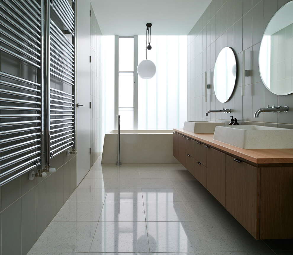 На фото: большая главная ванная комната в современном стиле с настольной раковиной, плоскими фасадами, столешницей из дерева, отдельно стоящей ванной, белой плиткой, каменной плиткой, серыми стенами, полом из травертина и темными деревянными фасадами с