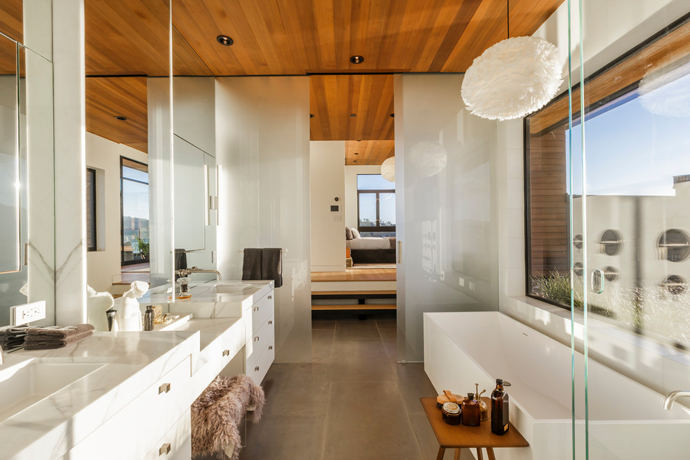 Cette photo montre une salle de bain principale tendance avec un placard à porte plane, des portes de placard blanches, une baignoire indépendante, un carrelage blanc, un mur blanc, un lavabo encastré, un sol gris et un plan de toilette blanc.