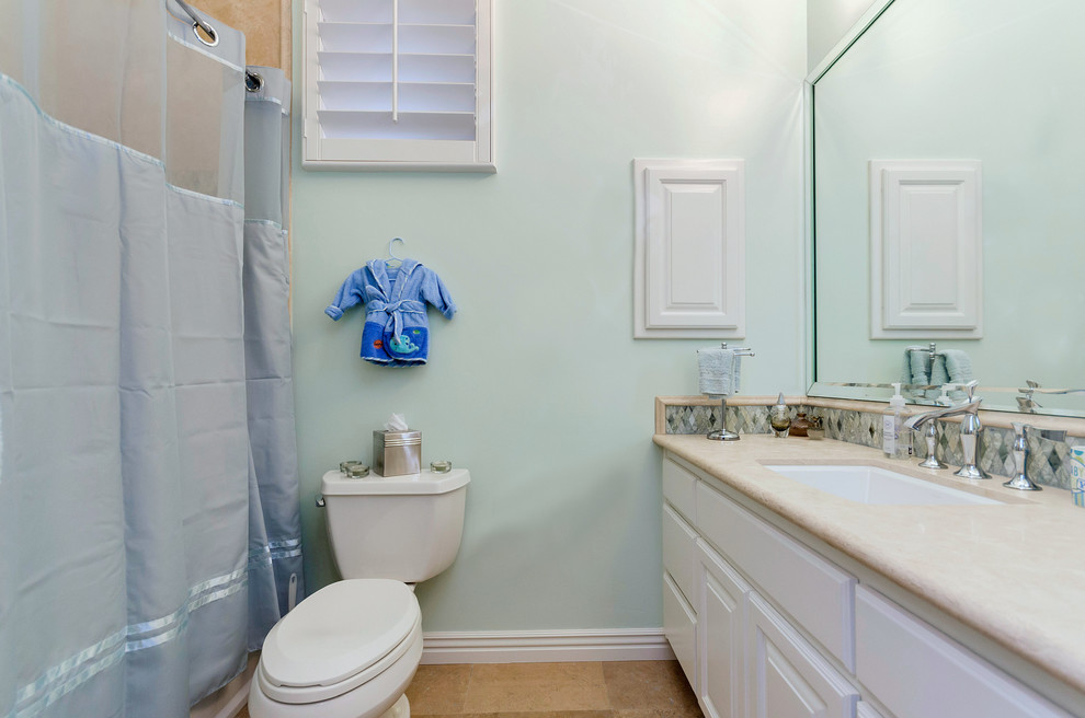 Klassisk inredning av ett badrum för barn, med ett undermonterad handfat, luckor med upphöjd panel, vita skåp, bänkskiva i kalksten, ett badkar i en alkov, en dusch/badkar-kombination, en toalettstol med hel cisternkåpa, gul kakel, glaskakel, gröna väggar och travertin golv
