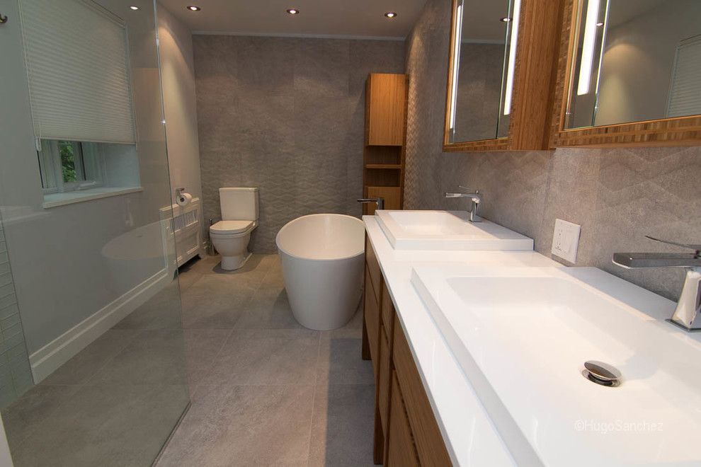 This is an example of a large contemporary ensuite bathroom in Montreal with a console sink, freestanding cabinets, medium wood cabinets, engineered stone worktops, a freestanding bath, a built-in shower, a one-piece toilet, grey tiles, porcelain tiles, grey walls and porcelain flooring.