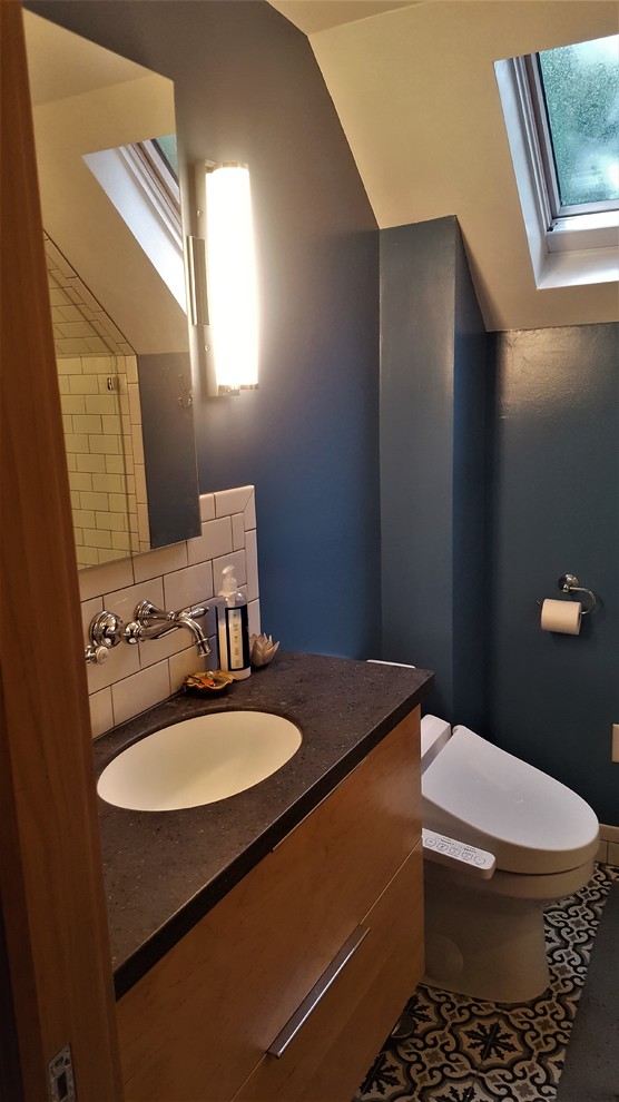 Foto på ett litet funkis en-suite badrum, med släta luckor, skåp i ljust trä, ett badkar i en alkov, en toalettstol med hel cisternkåpa, vit kakel, tunnelbanekakel, blå väggar, cementgolv, ett integrerad handfat, bänkskiva i akrylsten och flerfärgat golv