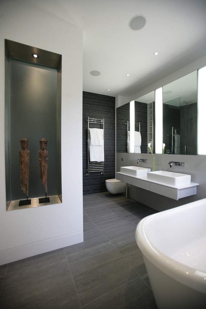 Photo of a medium sized contemporary ensuite bathroom in London with flat-panel cabinets, grey cabinets, a freestanding bath, a walk-in shower, a wall mounted toilet, grey tiles, porcelain tiles, grey walls, porcelain flooring and quartz worktops.