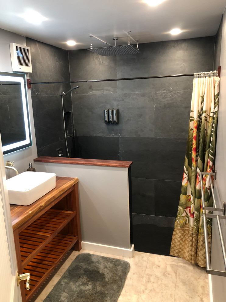Modern inredning av ett litet badrum med dusch, med skåp i mellenmörkt trä, en öppen dusch, en toalettstol med separat cisternkåpa, svart kakel, skifferkakel, ett fristående handfat och träbänkskiva