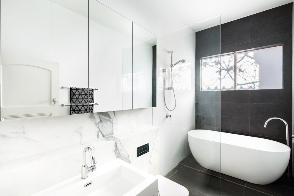 Photo of a medium sized contemporary family bathroom in Melbourne with glass-front cabinets, white cabinets, a freestanding bath, a walk-in shower, a wall mounted toilet, white tiles, marble tiles, white walls, ceramic flooring, an integrated sink, solid surface worktops, black floors and an open shower.