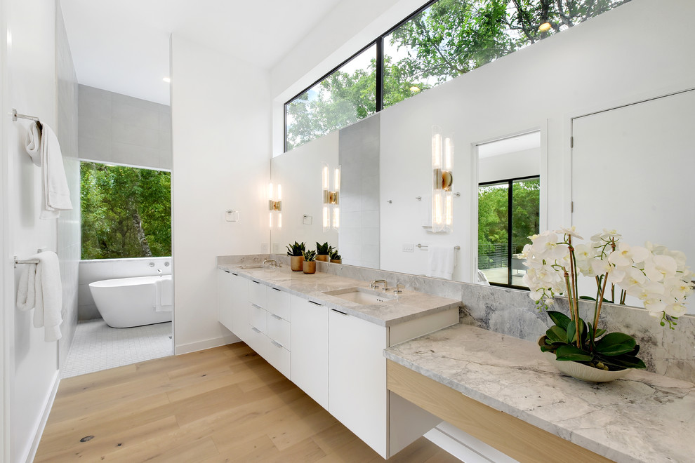 На фото: главная ванная комната в современном стиле с плоскими фасадами, белыми фасадами, отдельно стоящей ванной, серой плиткой, белыми стенами, светлым паркетным полом, врезной раковиной, бежевым полом, серой столешницей и зеркалом с подсветкой