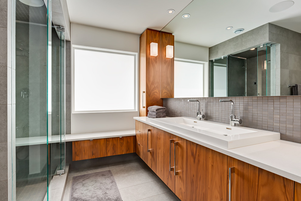 Modern inredning av ett badrum, med ett avlångt handfat, släta luckor, skåp i mellenmörkt trä, en dusch i en alkov, grå kakel, mosaik och grå väggar