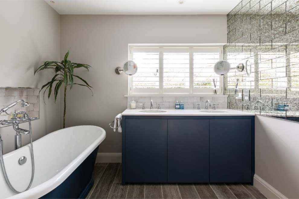 Medium sized contemporary ensuite bathroom in Berkshire with flat-panel cabinets, blue cabinets, a freestanding bath, porcelain flooring, grey floors, white worktops, solid surface worktops, beige walls, a submerged sink, grey tiles, glass tiles, a wall niche, double sinks and a built in vanity unit.