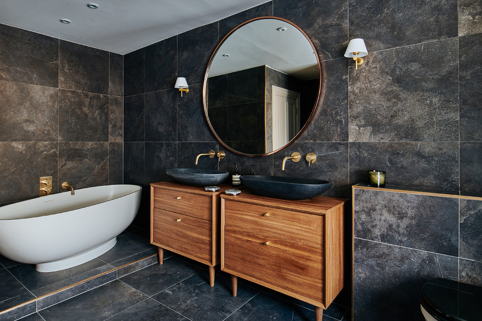 Diseño de cuarto de baño actual de tamaño medio con armarios con paneles lisos, puertas de armario de madera oscura, bañera exenta, baldosas y/o azulejos negros, lavabo sobreencimera, encimera de madera, suelo negro y encimeras marrones