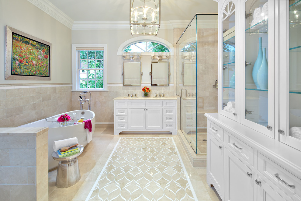 This is an example of a large classic ensuite bathroom in New York with recessed-panel cabinets, white cabinets, a freestanding bath, an alcove shower, a two-piece toilet, beige tiles, stone tiles, beige walls, a submerged sink, a hinged door and beige floors.