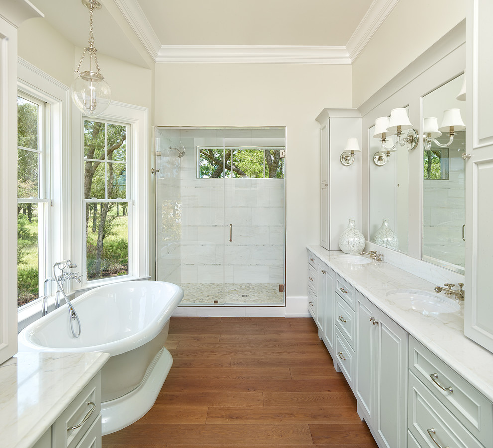 Bild på ett mellanstort vintage vit vitt en-suite badrum, med ett fristående badkar, mellanmörkt trägolv, marmorbänkskiva, en dusch i en alkov, beige väggar, ett undermonterad handfat, dusch med gångjärnsdörr, grå skåp, vit kakel, marmorkakel, brunt golv och luckor med infälld panel
