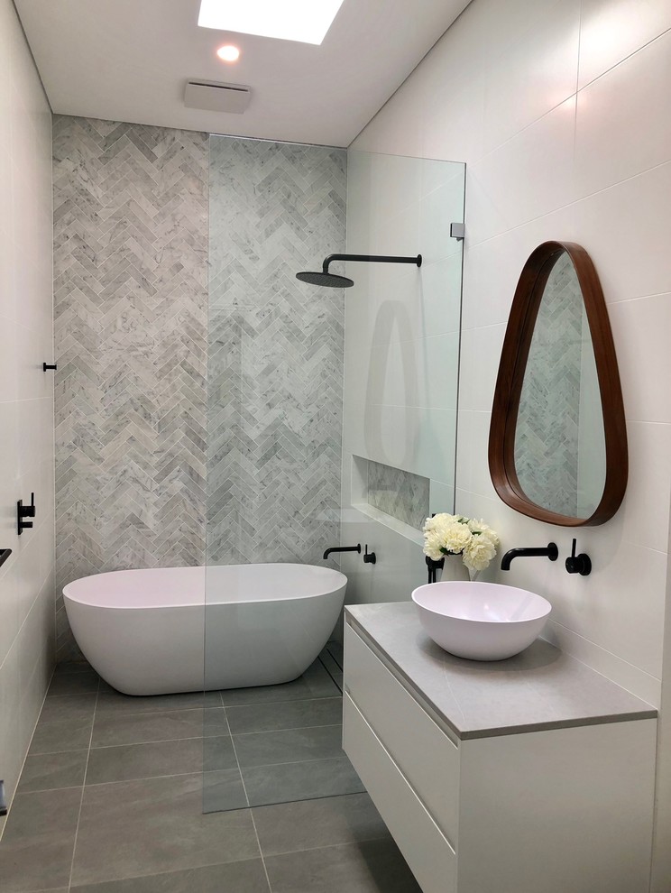Modern inredning av ett grå grått badrum, med släta luckor, vita skåp, ett fristående badkar, våtrum, grå kakel, ett fristående handfat, grått golv och med dusch som är öppen