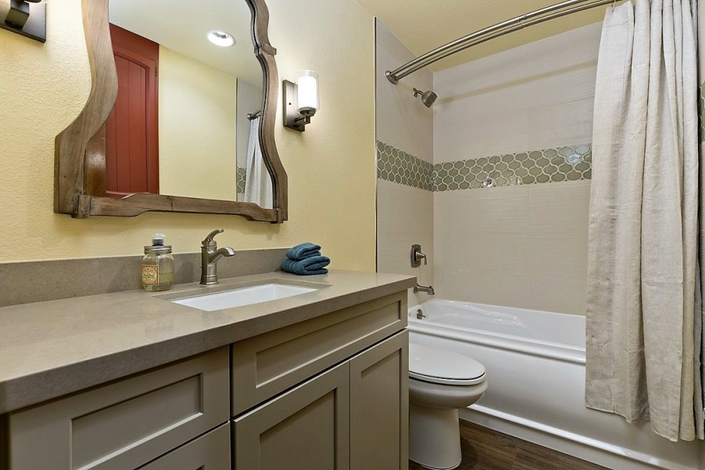 Klassisk inredning av ett litet brun brunt badrum, med skåp i shakerstil, gröna skåp, ett badkar i en alkov, en dusch/badkar-kombination, en toalettstol med hel cisternkåpa, flerfärgad kakel, gula väggar, laminatgolv, ett undermonterad handfat, bänkskiva i kvartsit, brunt golv och dusch med duschdraperi
