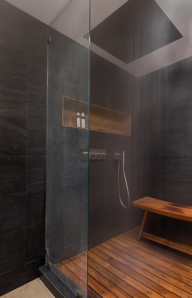 Photo of a large contemporary ensuite bathroom in New York with flat-panel cabinets, black cabinets, a freestanding bath, an alcove shower, a wall mounted toilet, black tiles, stone tiles, black walls, light hardwood flooring, an integrated sink, engineered stone worktops, beige floors and a hinged door.