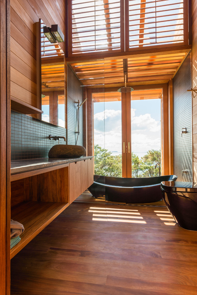 Cette image montre une salle de bain design en bois brun de taille moyenne avec une vasque, un placard à porte plane, un plan de toilette en surface solide, une baignoire indépendante, une douche ouverte, WC suspendus et un sol en bois brun.