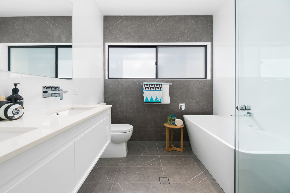 Cette image montre une salle de bain design avec un placard à porte plane, des portes de placard blanches, une baignoire indépendante, une douche d'angle, un carrelage gris, un carrelage blanc, un mur blanc, un lavabo encastré, un sol gris, une cabine de douche à porte battante et un plan de toilette beige.