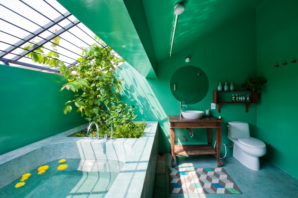 Exotisk inredning av ett badrum, med öppna hyllor, skåp i mörkt trä, en toalettstol med hel cisternkåpa, gröna väggar, betonggolv, ett fristående handfat, träbänkskiva och blått golv