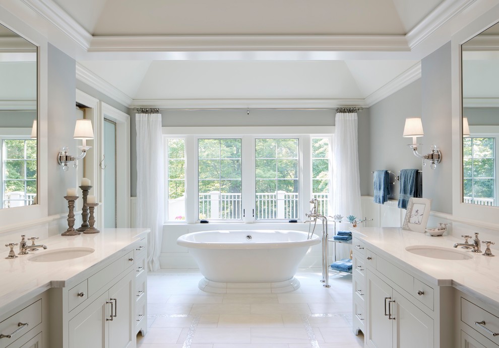 На фото: главная ванная комната в стиле неоклассика (современная классика) с фасадами с утопленной филенкой, белыми фасадами, отдельно стоящей ванной, серыми стенами, врезной раковиной, белым полом и белой столешницей