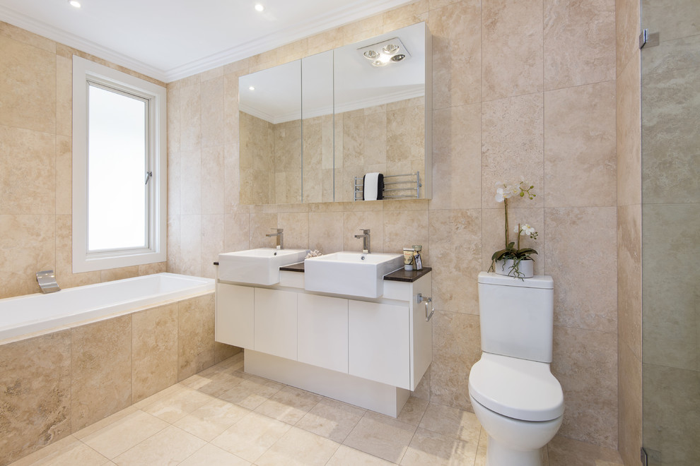 Modernes Badezimmer mit flächenbündigen Schrankfronten, weißen Schränken, Badewanne in Nische, Duschnische, Wandtoilette mit Spülkasten, beiger Wandfarbe, Kalkstein und Kalkfliesen in Sydney