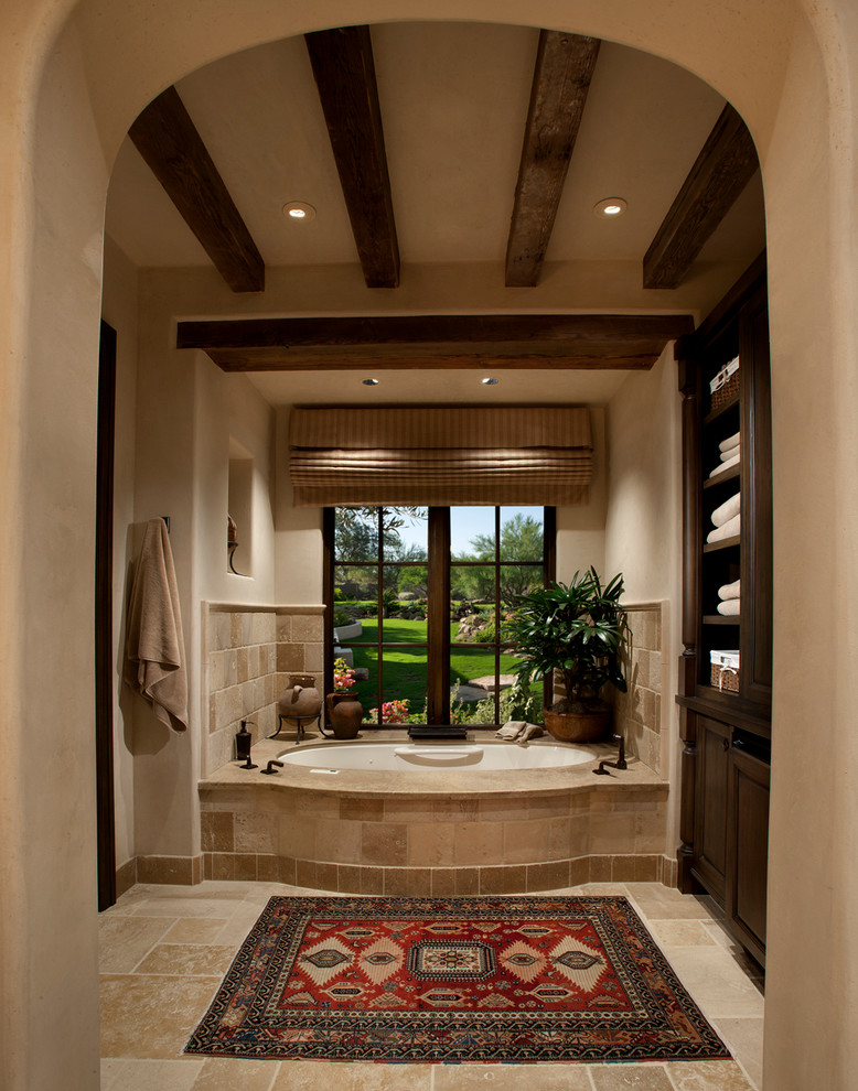 На фото: главная ванная комната среднего размера в средиземноморском стиле с фасадами с выступающей филенкой, темными деревянными фасадами, бежевой плиткой, каменной плиткой, полновстраиваемой ванной, бежевыми стенами и полом из известняка с