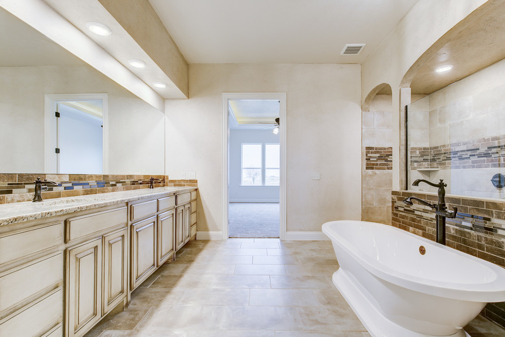 Пример оригинального дизайна: большая главная ванная комната в средиземноморском стиле с фасадами с выступающей филенкой, искусственно-состаренными фасадами, отдельно стоящей ванной, двойным душем, коричневой плиткой, удлиненной плиткой, бежевыми стенами, врезной раковиной, столешницей из гранита, серым полом, открытым душем и бежевой столешницей