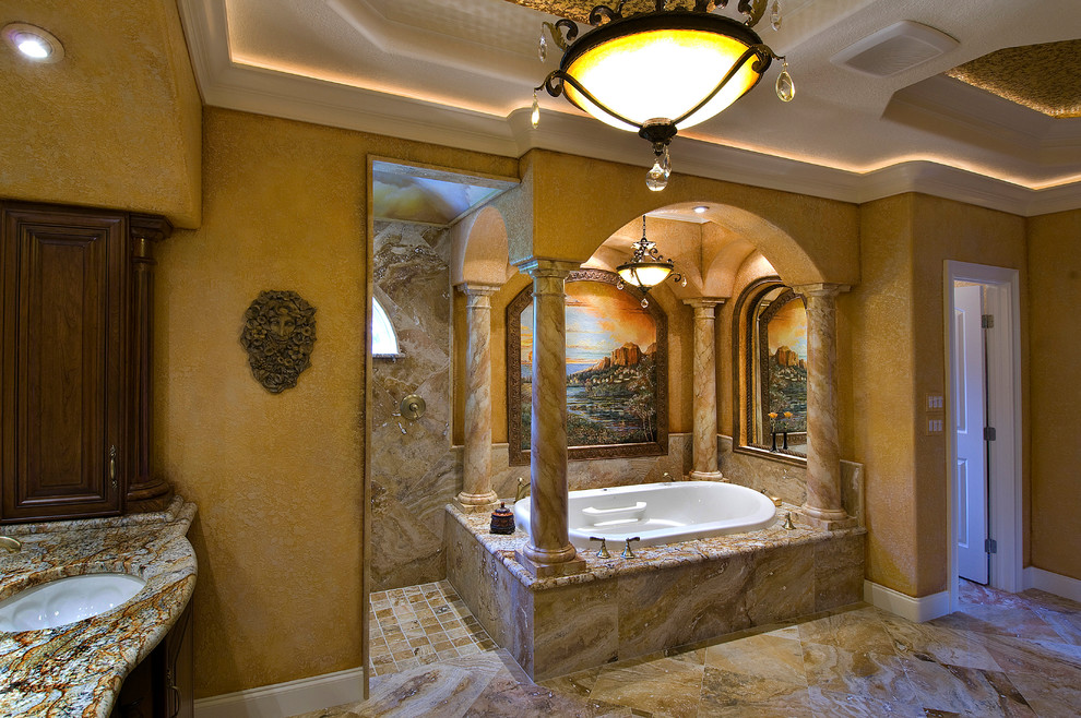 Cette photo montre une très grande salle de bain principale méditerranéenne en bois foncé avec un placard avec porte à panneau surélevé, une baignoire posée, un carrelage beige, des dalles de pierre, un mur beige, un sol en ardoise, un lavabo encastré, un plan de toilette en quartz, un sol beige et un plan de toilette beige.