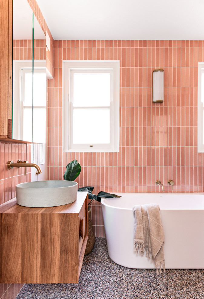 На фото: главная ванная комната среднего размера в скандинавском стиле с отдельно стоящей ванной, открытым душем, раздельным унитазом, розовой плиткой, керамической плиткой, розовыми стенами, полом из терраццо, подвесной раковиной, разноцветным полом, открытым душем, тумбой под одну раковину, подвесной тумбой, фасадами цвета дерева среднего тона, столешницей из дерева и коричневой столешницей с
