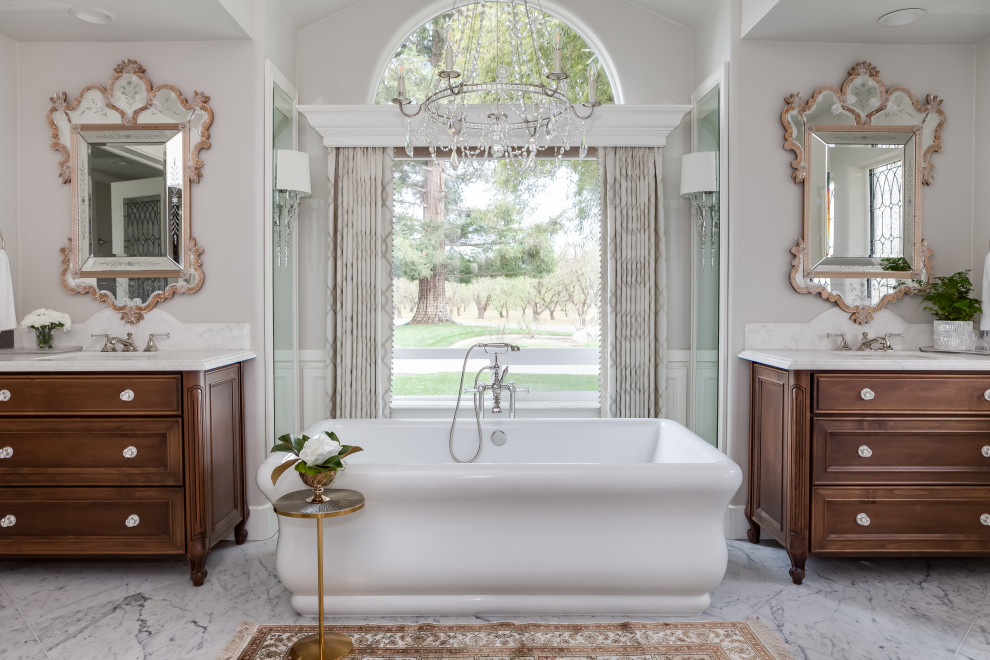 Inredning av ett klassiskt stort vit vitt en-suite badrum, med skåp i mellenmörkt trä, ett fristående badkar, en öppen dusch, en toalettstol med hel cisternkåpa, vit kakel, stenhäll, grå väggar, marmorgolv, ett undermonterad handfat, marmorbänkskiva, vitt golv, med dusch som är öppen och luckor med infälld panel