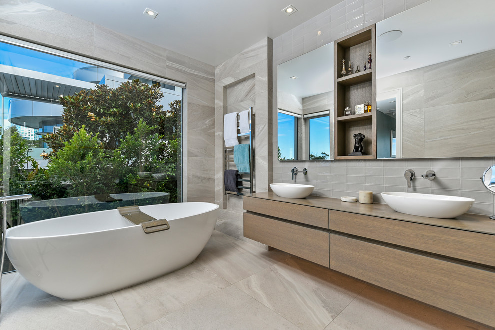 Bild på ett funkis beige beige badrum, med släta luckor, skåp i ljust trä, ett fristående badkar, grå kakel, grå väggar, ett fristående handfat, träbänkskiva och grått golv
