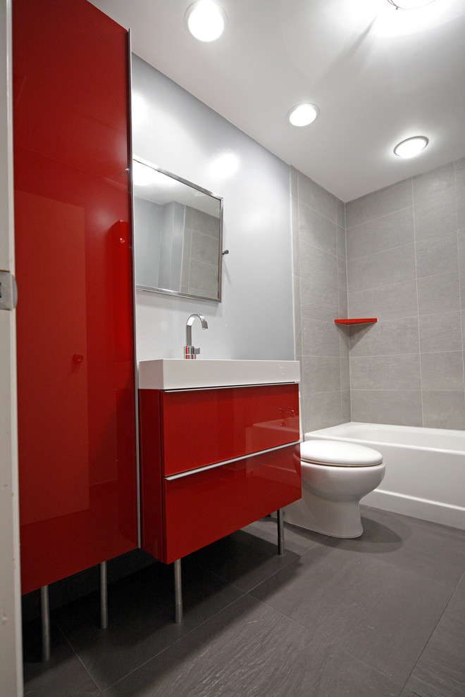 Cette image montre une salle de bain minimaliste de taille moyenne pour enfant avec un placard à porte plane, des portes de placard rouges, une baignoire en alcôve, un combiné douche/baignoire, WC séparés, un carrelage gris, des carreaux de porcelaine, un mur bleu, un sol en carrelage de porcelaine, un lavabo intégré et un plan de toilette en surface solide.