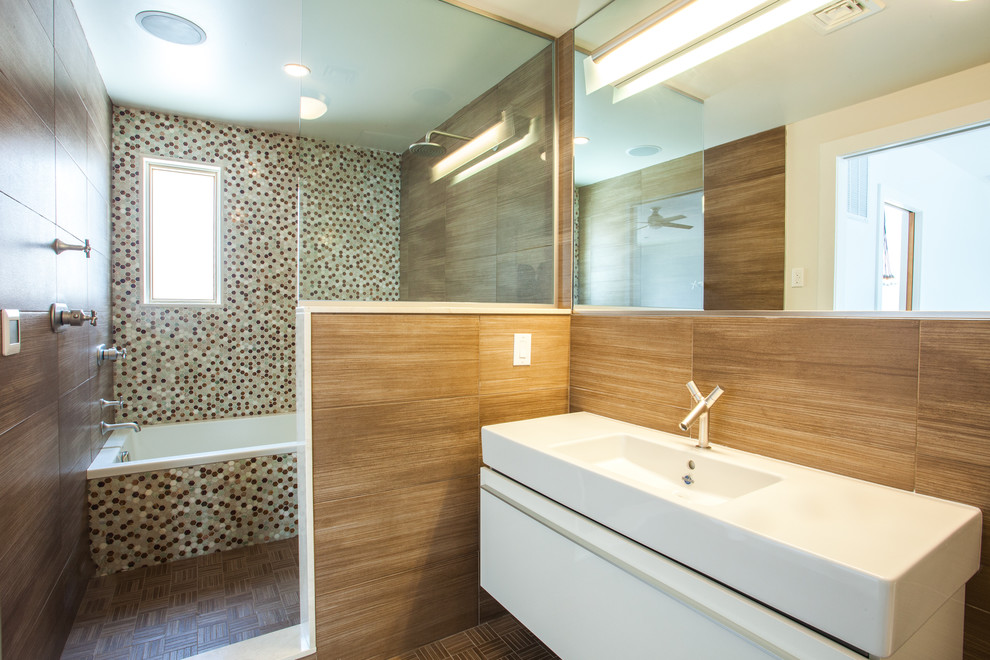 Modelo de cuarto de baño contemporáneo con lavabo integrado, armarios con paneles lisos, puertas de armario blancas, encimera de acrílico, bañera empotrada, combinación de ducha y bañera, sanitario de una pieza, baldosas y/o azulejos marrones, imitación madera y ventanas