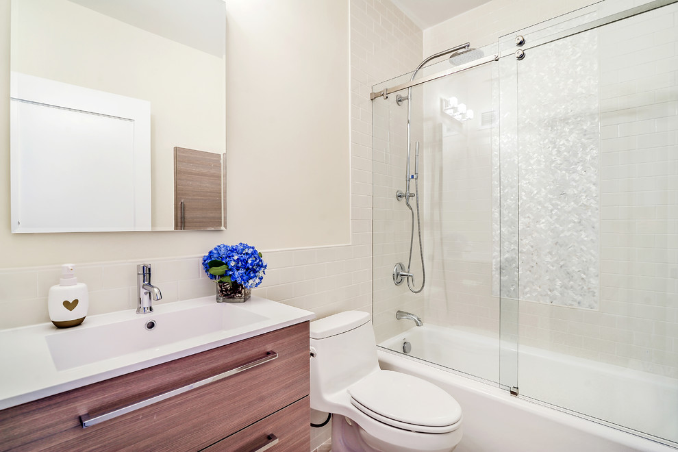 Photo of a medium sized classic shower room bathroom in New York with flat-panel cabinets, medium wood cabinets, an alcove bath, a shower/bath combination, a one-piece toilet, solid surface worktops, white tiles, metro tiles, white walls and an integrated sink.