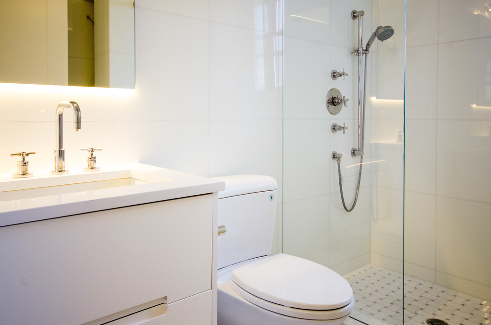 Modern inredning av ett litet badrum med dusch, med ett nedsänkt handfat, släta luckor, vita skåp, bänkskiva i akrylsten, en dusch i en alkov, en toalettstol med hel cisternkåpa, vit kakel, glaskakel, vita väggar och klinkergolv i keramik