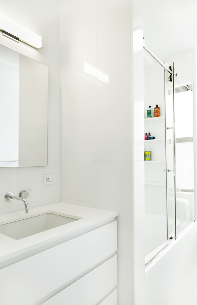 This is an example of a small contemporary family bathroom in New York with white cabinets, a built-in bath, a shower/bath combination, a one-piece toilet, white tiles, porcelain tiles, white walls, a submerged sink, solid surface worktops, flat-panel cabinets and porcelain flooring.