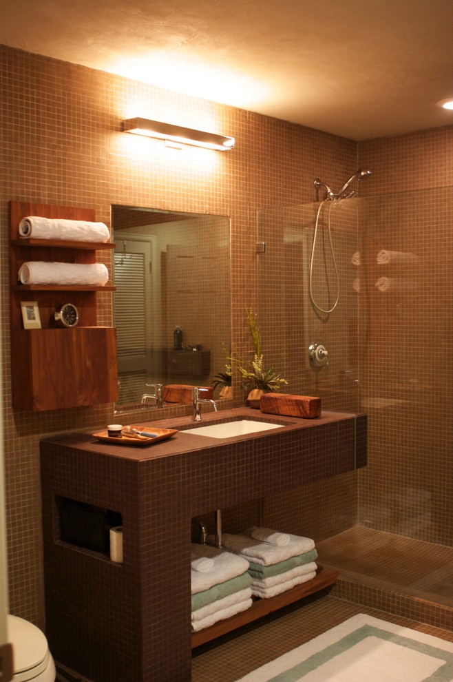 Modern inredning av ett litet brun brunt badrum med dusch, med en öppen dusch, brun kakel, klinkergolv i keramik, ett undermonterad handfat, kaklad bänkskiva, öppna hyllor, bruna skåp, en toalettstol med hel cisternkåpa, keramikplattor, bruna väggar, brunt golv och med dusch som är öppen