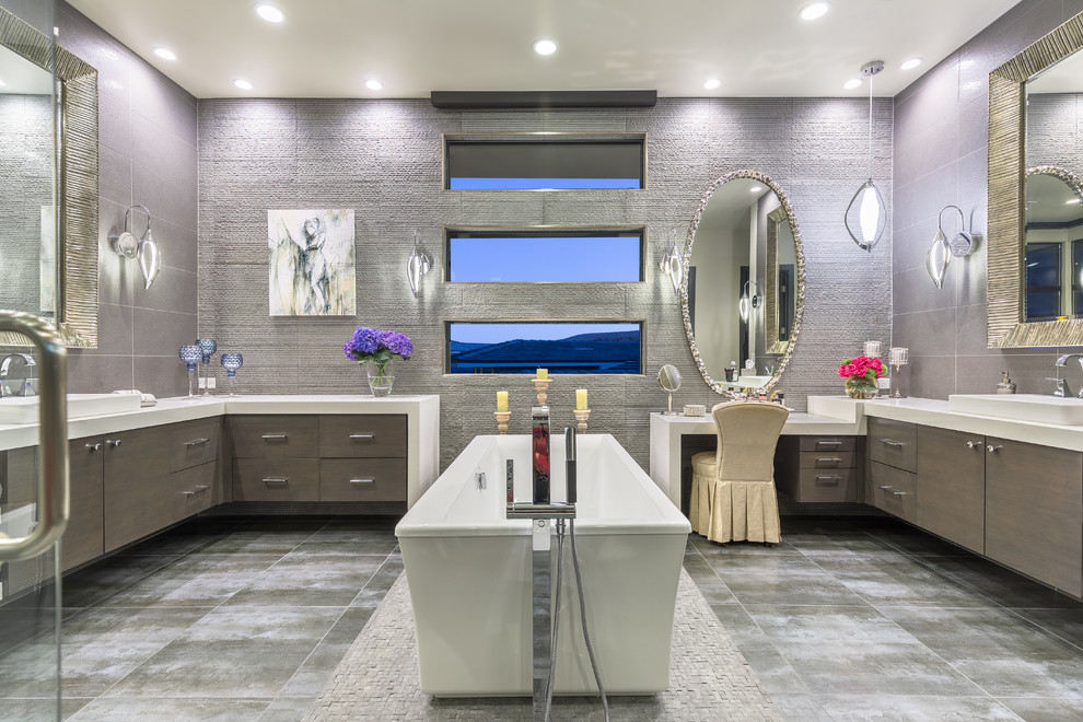 Cette image montre une salle de bain design avec un placard à porte plane, des portes de placard grises, une baignoire indépendante, un carrelage gris, une vasque, un sol gris et un plan de toilette blanc.
