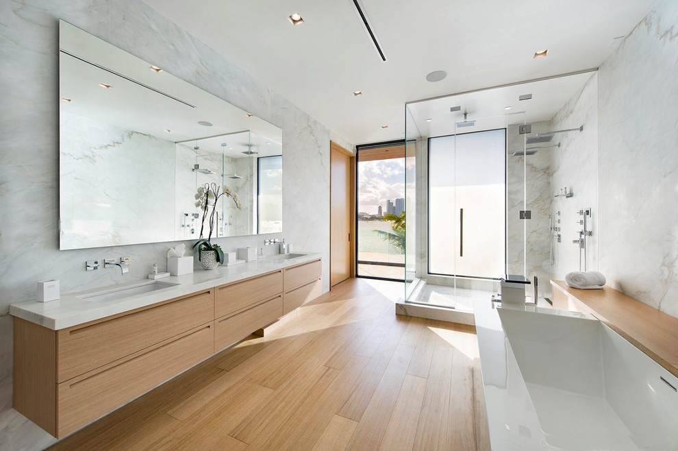 Inspiration for a large contemporary ensuite bathroom in Miami with flat-panel cabinets, light wood cabinets, a freestanding bath, a double shower, white tiles, stone slabs, white walls, light hardwood flooring, a submerged sink, a hinged door, white worktops, a one-piece toilet, marble worktops and beige floors.
