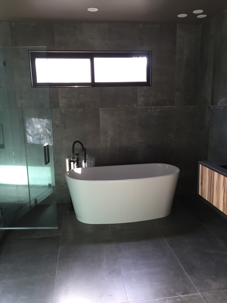 Modern inredning av ett stort en-suite badrum, med släta luckor, skåp i ljust trä, ett fristående badkar, en dusch i en alkov, grå kakel, cementkakel, grå väggar, cementgolv, ett undermonterad handfat, bänkskiva i akrylsten, grått golv och dusch med gångjärnsdörr