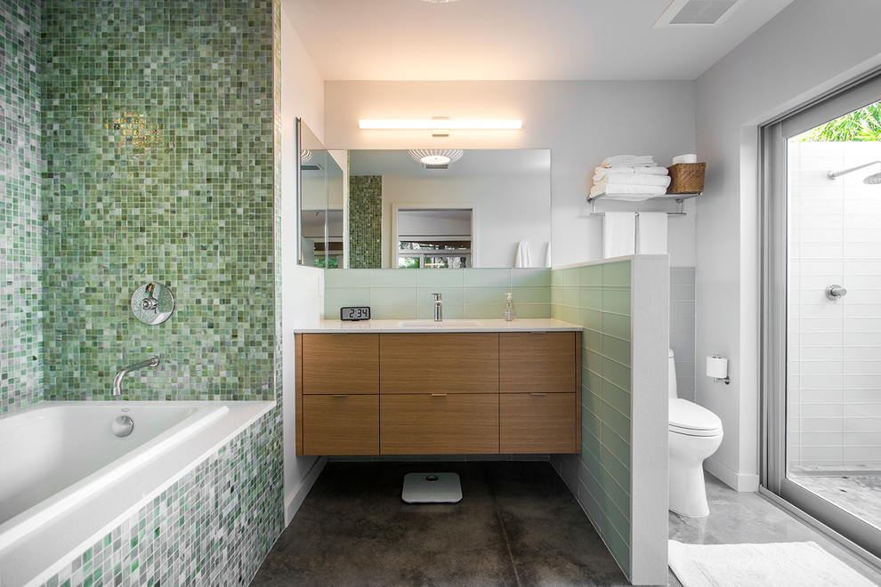 タンパにあるミッドセンチュリースタイルのおしゃれな浴室 (フラットパネル扉のキャビネット、緑のタイル、モザイクタイル、ドロップイン型浴槽、オープン型シャワー、一体型シンク、白い壁、コンクリートの床、中間色木目調キャビネット) の写真
