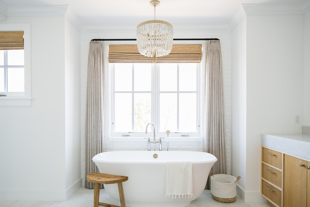 Diseño de cuarto de baño principal costero con armarios con paneles lisos, puertas de armario de madera clara, bañera exenta y paredes blancas