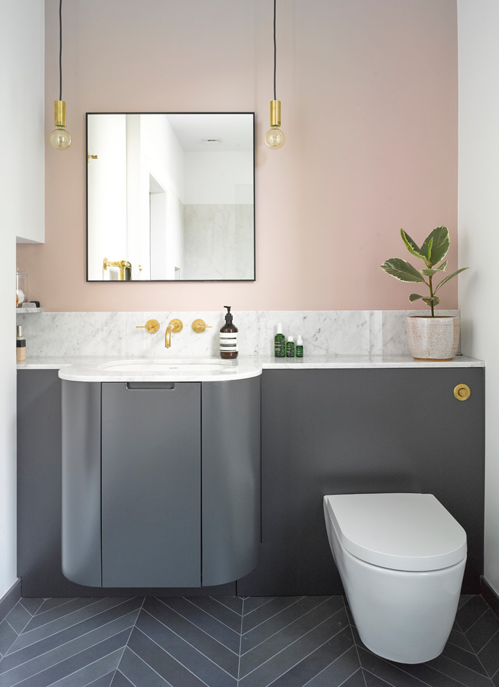Modern inredning av ett badrum, med släta luckor, grå skåp, en vägghängd toalettstol, rosa väggar, ett undermonterad handfat, grått golv, vit kakel, marmorkakel och marmorbänkskiva