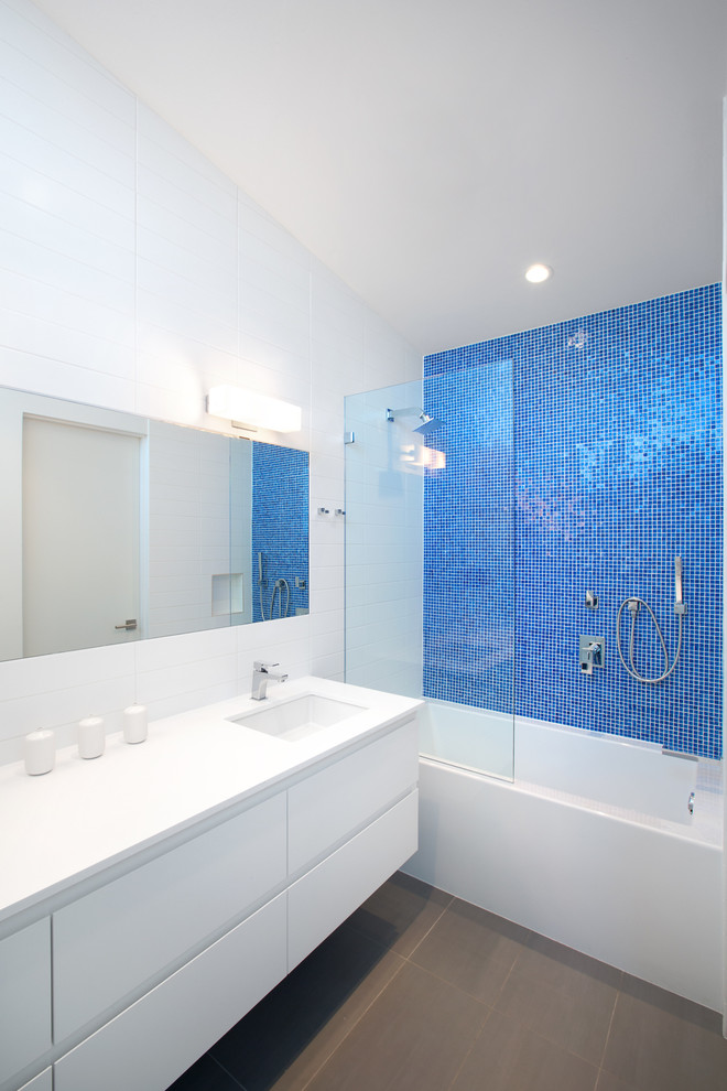 Inredning av ett modernt badrum, med ett undermonterad handfat, släta luckor, vita skåp, bänkskiva i kvartsit, en vägghängd toalettstol, blå kakel, mosaik, vita väggar, klinkergolv i porslin, ett badkar i en alkov och en dusch/badkar-kombination