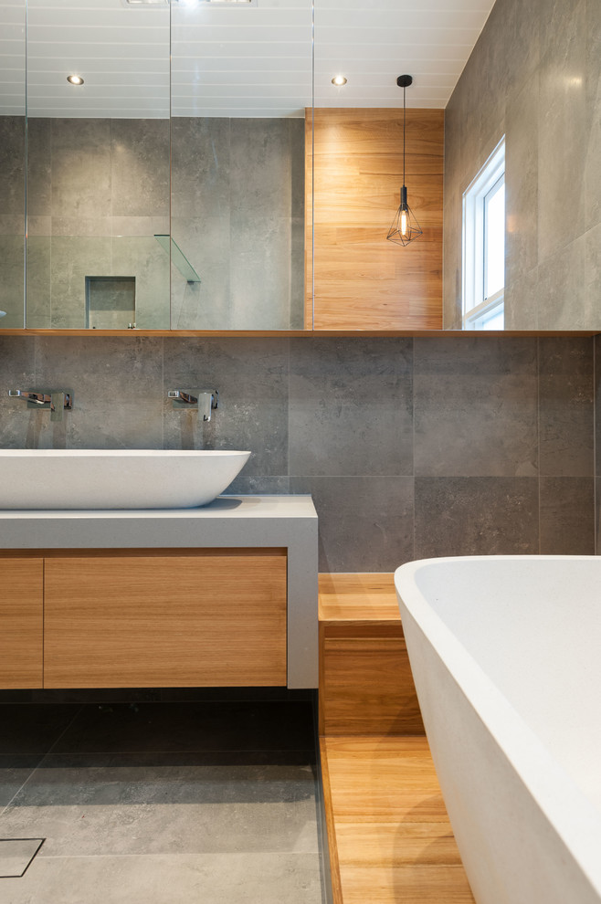 Пример оригинального дизайна: большая главная ванная комната в современном стиле с светлыми деревянными фасадами, отдельно стоящей ванной, открытым душем, серой плиткой, керамической плиткой, серыми стенами, полом из керамической плитки, консольной раковиной и столешницей из искусственного кварца