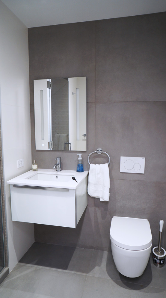 Bild på ett stort funkis vit vitt en-suite badrum, med släta luckor, vita skåp, en dusch i en alkov, en vägghängd toalettstol, grå kakel, porslinskakel, grå väggar, klinkergolv i porslin, ett väggmonterat handfat, bänkskiva i akrylsten, grått golv och dusch med gångjärnsdörr