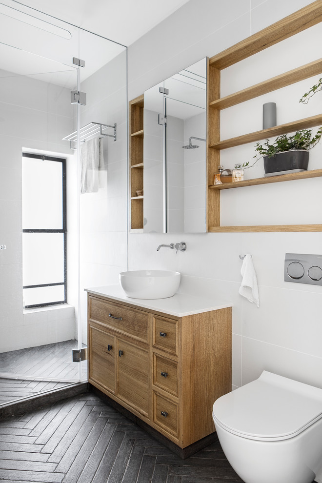 Foto på ett minimalistiskt vit badrum, med skåp i mellenmörkt trä, en dusch i en alkov, en vägghängd toalettstol, vit kakel, vita väggar, ett fristående handfat, grått golv, dusch med gångjärnsdörr och luckor med infälld panel