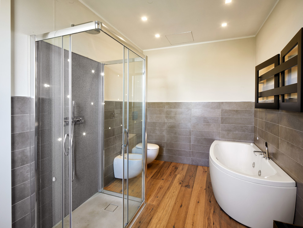 На фото: главная ванная комната в современном стиле с душем без бортиков, серой плиткой, душем с раздвижными дверями, угловой ванной, биде, паркетным полом среднего тона, коричневым полом и серыми стенами